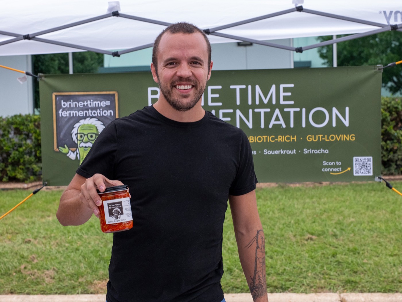 A vendor holding a jar of garlic ginger kimchi in front of a Brine and Time Fermentation banner